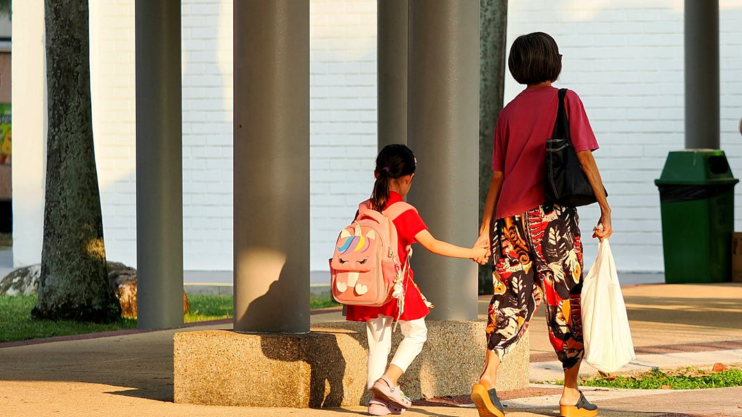 A child with a pink school bag holding an adult's hand on the walk to school, seen from behind