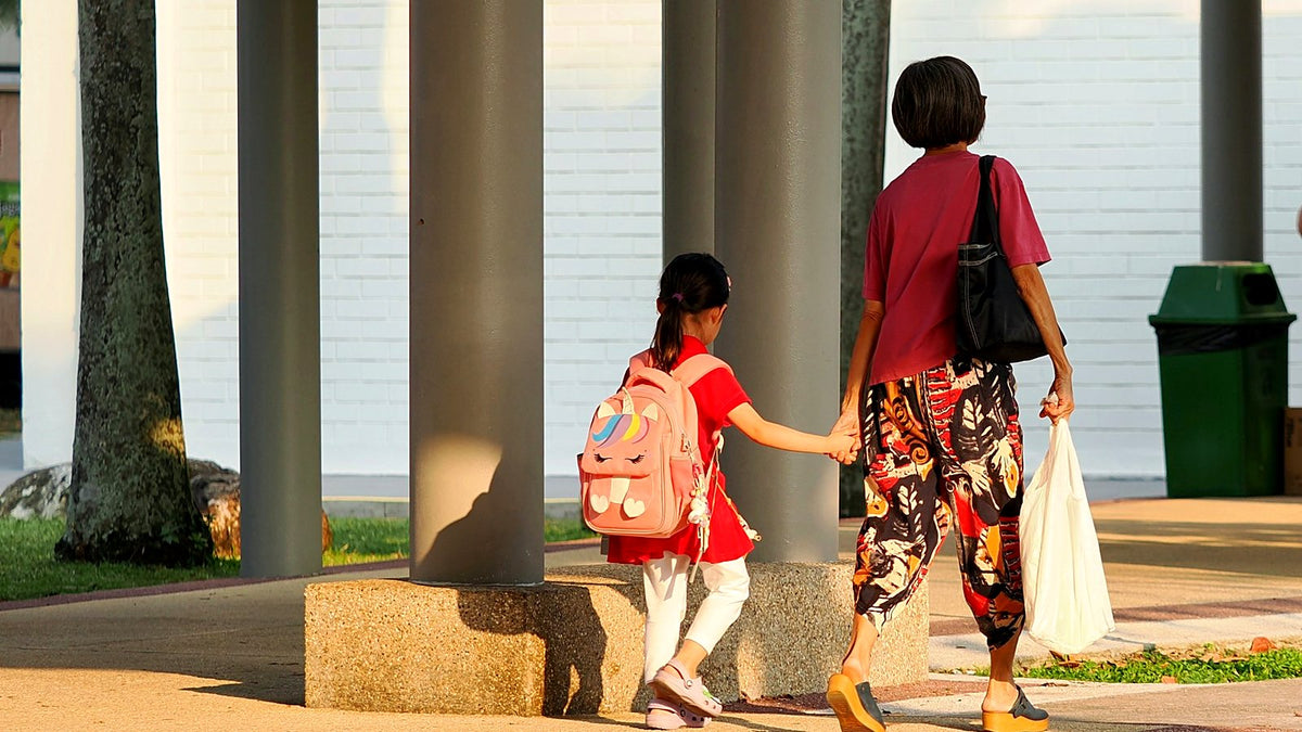 A child with a pink school bag holding an adult's hand on the walk to school, seen from behind